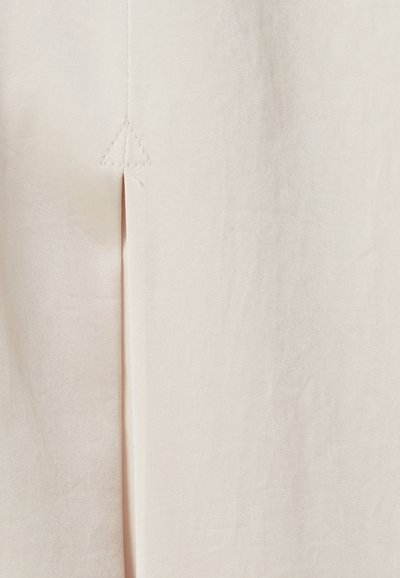 Primo piano di un tessuto beige con una cucitura verticale e un piccolo dettaglio cucito a forma di triangolo sopra uno spacco.