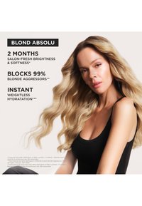 Femme blonde aux longs cheveux ondulés portant un débardeur noir, faisant la promotion des bienfaits d'un produit capillaire pour la brillance, la douceur et l'hydratation.