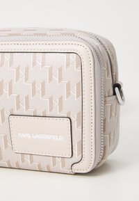 Krem i bež crossbody torba s utisnutim logotipom, koja se odlikuje glatkom kožnom teksturom, srebrnim hardverom i zatvaranjem na zatvarač.