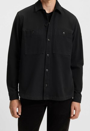 Mand iført sort skjorte med knapper og to brystlommer med klapper samt sorte bukser, stående mod en ensartet lys baggrund.