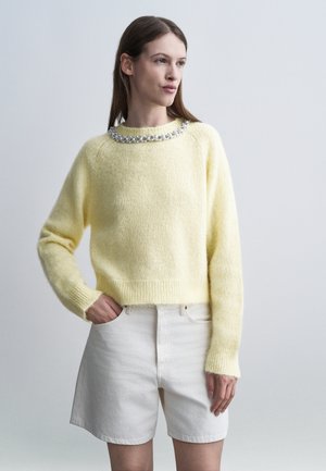 Femme portant un pull léger jaune pâle en tissu duveteux avec des perles décoratives au col et un short blanc taille haute, debout avec une main dans la poche.