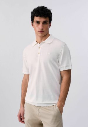 Giovane uomo con capelli ricci e scuri che indossa una maglietta polo bianca a maniche corte e pantaloni beige su uno sfondo chiaro e uniforme.