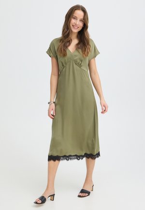 Žena sa usmieva, má na sebe olivovo zelené midi šaty s výstrihom do V, krátkymi rukávmi, čiernym čipkovým lemom a čiernymi sandálmi na opätku, stojí na bielom pozadí.