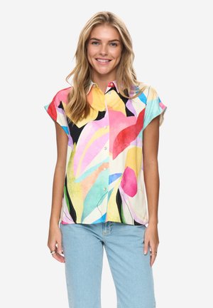 Femme souriante portant une chemise à manches courtes à boutons avec un imprimé abstrait coloré et un jean bleu clair, sur un fond uni.