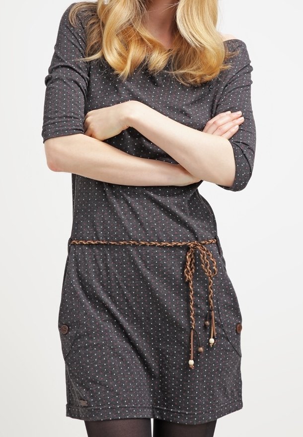 Femme aux cheveux blonds portant une robe gris foncé à pois avec des manches trois-quarts, une ceinture tressée et les bras croisés.