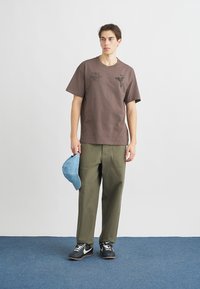 Brun bomullst-shirt med grafik och text, kombinerad med olivgröna byxor. Modellen håller en ljusblå keps. Svarta och vita sneakers.