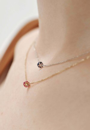 Collares de oro y plata superpuestos sobre la piel; el de oro cuenta con una gema rosa, el de plata con una gema azul, ambos con cadenas delicadas.