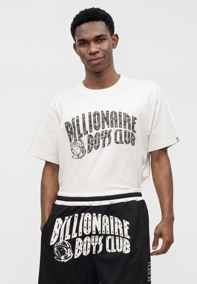 Νέος άνδρας που φοράει μια λευκή μπλούζα Billionaire Boys Club και μαύρα σορτς με το ίδιο λογότυπο, στέκεται μπροστά σε ένα απλό φωτεινό φόντο.