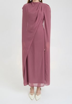 Femme portant une longue robe mauve avec un devant drapé et des manches transparentes, associée à des chaussures blanches pointues ornées de détails en perles.