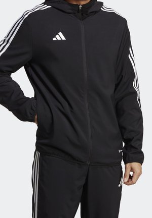 Mand iført sort Adidas træningsdragt med hvide striber på ærmer og bukser, hænder delvist i lommerne, stående mod ensfarvet baggrund.