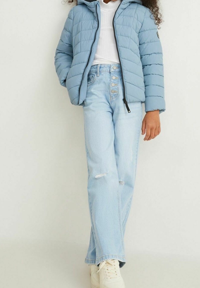 Jeune personne portant une doudoune bleu clair, un col roulé blanc, un jean taille haute bleu clair avec une braguette à boutons et des baskets blanches.