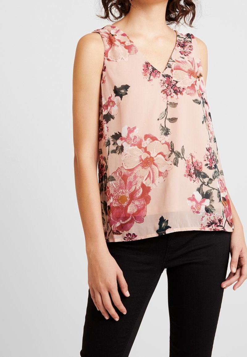Blouse sans manches rose à motif floral avec col en V portée avec un pantalon noir sur une personne aux cheveux bouclés et fond neutre.