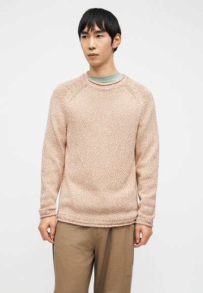 Homme aux cheveux noirs courts portant un pull beige texturé et un pantalon marron clair sur un fond blanc uni.