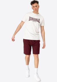Hvid t-shirt med maroon "Lonsdale London" logo, parret med maroon shorts. Modellen bærer hvide sneakers og hvide strømper.