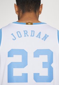 Maillot de basket-ball blanc avec des accents bleu clair. Présente le nom « JORDAN » et le numéro « 23 » en lettres brodées bleu clair. Tissu à texture en maille.