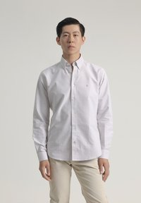Camisa blanca de manga larga y botones con finas rayas verticales azules, que presenta un cuello puntiagudo y un pequeño logo en el lado izquierdo del pecho. Pantalones en tonos neutros.