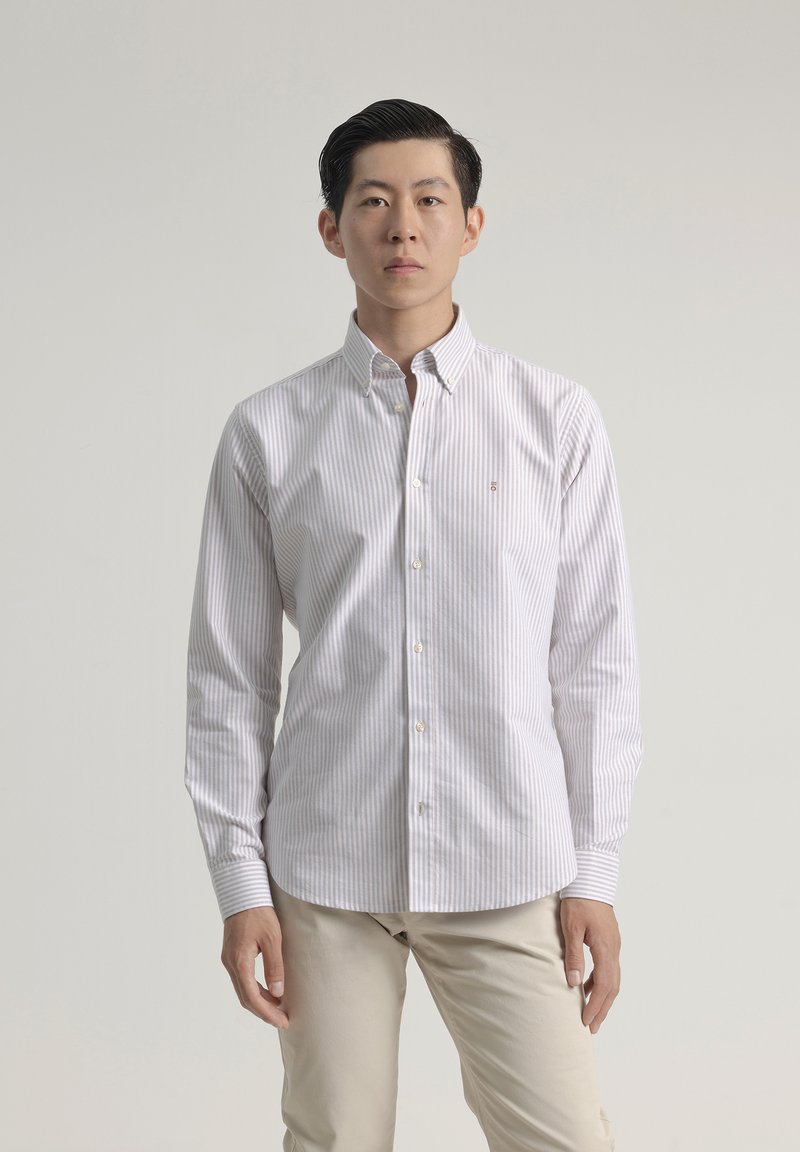 Camisa blanca de manga larga y botones con finas rayas verticales azules, que presenta un cuello puntiagudo y un pequeño logo en el lado izquierdo del pecho. Pantalones en tonos neutros.