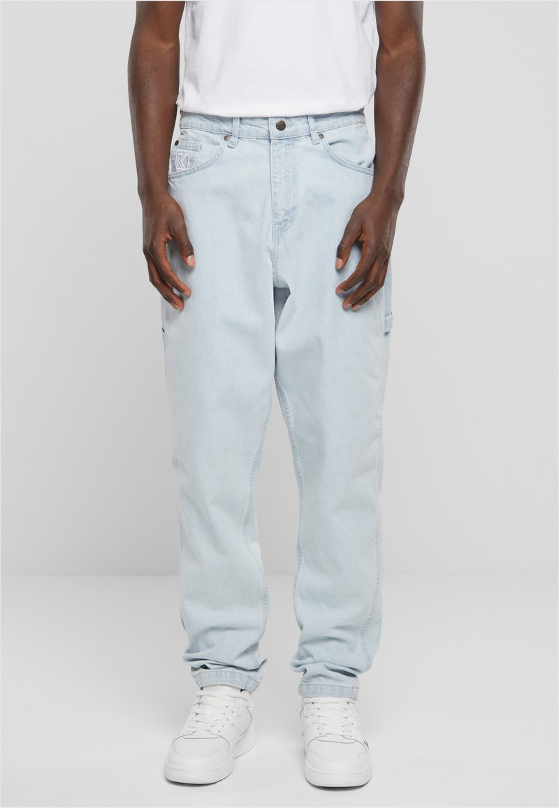 Light blue denim pants with a loose fit and tapered cuffs, featuring a front button closure and side pockets. Worn with white sneakers.