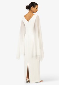 White fitted dress with a V-back, long sheer sleeves, back zipper, and a high slit at the hem. Smooth fabric with elegant draping accents.