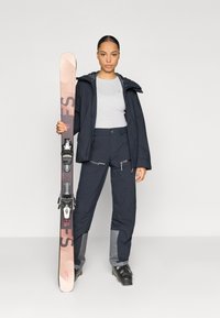 Costum de schi format dintr-o jachetă impermeabilă albastru închis cu glugă, bluză termică gri, pantaloni închiși la culoare cu fermoare și cizme de schi negre. Țin schiurile în mână.