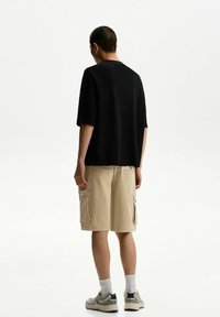 Tricou negru cu mâneci scurte și croială lejeră, asortat cu pantaloni scurți cargo bej, prevăzuți cu buzunare și textură uniformă. Adidași gri cu accente.