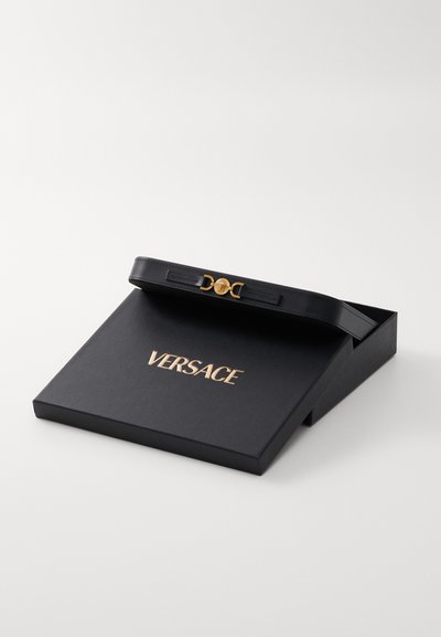 Caja negra de Versace con logo dorado, con una correa de cuero negro y un acento de hardware dorado circular en la parte superior. Diseño simple y elegante.
