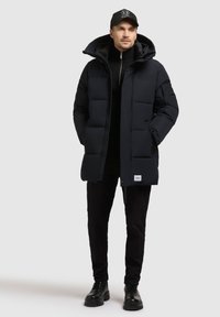 Jachetă puffer neagră cu umplutură, cu guler înalt și glugă detașabilă, având un fermoar frontal și un logo brodat în partea de jos. Pantaloni negri și bocanci.
