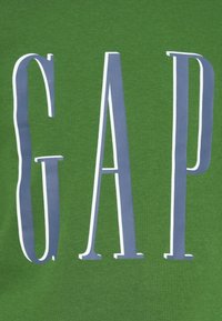 Πράσινη βαμβακερή μπλούζα με bold, κατακόρυφα γράμματα που γράφουν "GAP" σε μπλε χρώμα με λευκά περιγράμματα, πάνω σε μαλακό, υφασμάτινο ύφασμα.