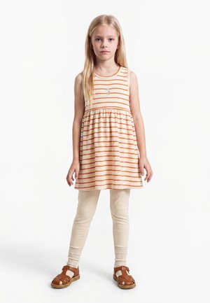 Jeune fille aux longs cheveux blonds portant une robe sans manches rayée crème et orange, des leggings crème et des sandales marron, debout sur un fond blanc.