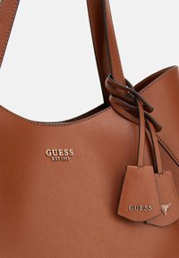 Braune strukturierte Tragetasche mit eingeprägtem "GUESS EST 1981" Logo, zwei Akzent-Tags und glatten Ledergriffen.
