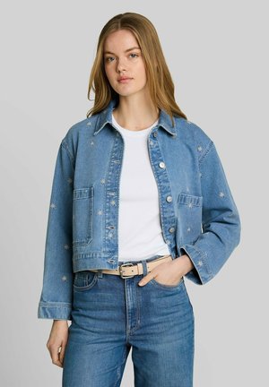 Junge Frau mit langen Haaren trägt eine blaue Jeansjacke mit Sternstickerei, weißes Hemd, blaue Jeans und beigen Gürtel, vor einem grauen Hintergrund stehend.