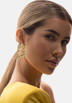 Kobieta o blond włosach zaczesanych do tyłu, ubrana w żółtą bluzkę i duże, teksturowane złote kolczyki-koła, spoglądająca przez ramię.