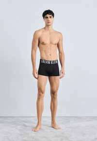 Crne pamučne bokserice s širokim elastičnim pojasom na kojem je istaknut logo "CALVIN KLEIN". Model stoji bos na svijetlosivoj tepisonu.