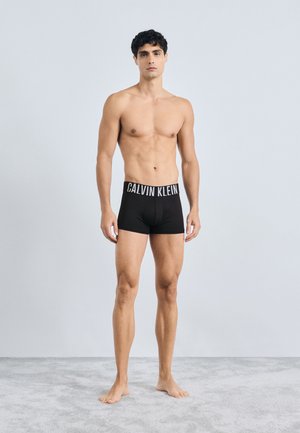 Boxers de algodão pretos com uma faixa elástica larga com o logótipo "CALVIN KLEIN". Modelo está descalço numa superfície carpetada cinza clara.