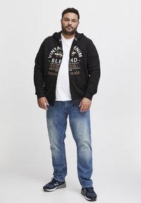 Crna zip-up dukserica s dizajnom teksta, bijela majica ispod, svijetloplave traperice i crne atletske tenisice, stojeći protiv jednostavne pozadine.