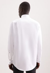 Camicia bianca realizzata in tessuto liscio, con colletto alto, maniche lunghe e polsini abbottonati con una leggera curvatura all'orlo.