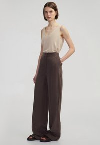 Beige bezrukávový top s výstřihem do V, spárovaný s hnědými kalhotami s vysokým pasem a širokými nohavicemi. Hladká textilie poskytující uvolněný siluetu.