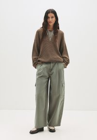 Brun struktureret oversized sweater, olivengrøn cargo-bukser med sidelommer og mørke sko med et mønstrede accent. Model står foran en hvid baggrund.