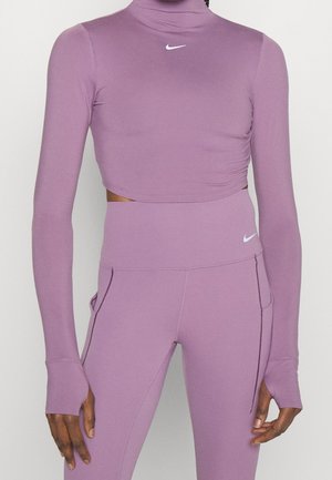 Kobieta ubrana w jasnofioletowy krótki top z długim rękawem oraz legginsy z wysokim stanem z logo Nike, stojąca na jednolitym tle.