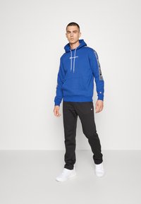 Felpa con cappuccio blu con cordini, tasca frontale e logo bianco, abbinata a pantaloni neri e sneaker bianche, in piedi su uno sfondo semplice.
