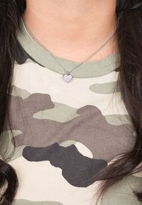 Zilveren ketting met een hartvormige hanger voorzien van roze edelsteenaccenten, gedragen over een camouflagepatroonkleurig shirt.