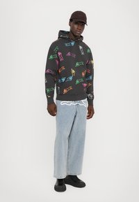 Moletom preto com designs de animais coloridos e contornados, combinado com jeans largos azul claro e sapatos pretos. Boné simples usado na cabeça.