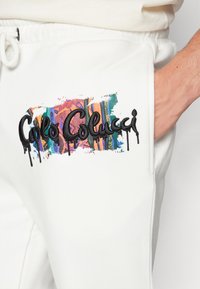 Hvite joggebukser med en fargerik, mønstret lapp som har brodert teksten "Colo Colucci," komplettert med svarte drip-detaljer.