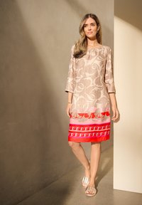 Sukienka w kwiatowy wzór w kolorze beżowym, z jasnoczerwoną i różową lamówką w geometryczne akcenty. Krótkie rękawy i okrągły dekolt.