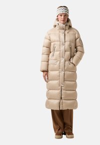 Beige pufferjack met een lange, gewatteerde design, ritssluiting aan de voorkant en meerdere zakken. Draagt het met bruine broek en een gestreepte hoofdband.