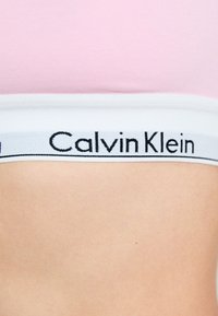 Světle růžový bavlněný bralet s bílým elastickým páskem, na kterém je černý brand "Calvin Klein". Detailní pohled na pásek a texturu látky.