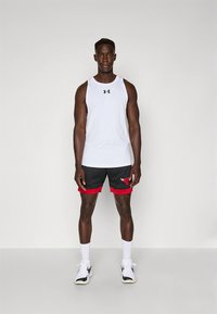 Camisola desportiva branca sem mangas, calções pretos com acabamento vermelho e um logótipo. Meias brancas e sapatos desportivos pretos completam o conjunto.