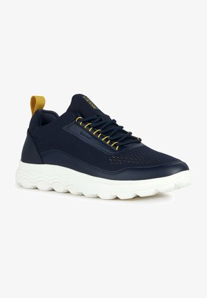 Zapatillas de deporte navy con parte superior de tela texturizada, cordones amarillos y suela de goma blanca con patrones de olas. Tirador en la parte trasera.