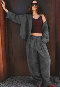 Femme aux cheveux bouclés portant un costume gris oversize, un crop top bordeaux et des lunettes de soleil, appuyée contre une porte en métal couleur rouille.