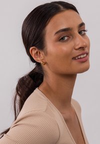 Goldene Creolen, die von einem Modell getragen werden, mit einer glatten, glänzenden Oberfläche. Das Modell hat eine mittelbraune Hautfarbe und lange, dunkle Haare.